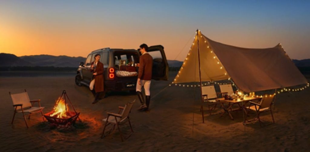 Un couple prépare un bivouac de luxe sur une plage désertique à côté d'un SUV iCAUR tout-terrain au coucher du soleil, avec un feu de camp et une tente éclairée par des guirlandes.