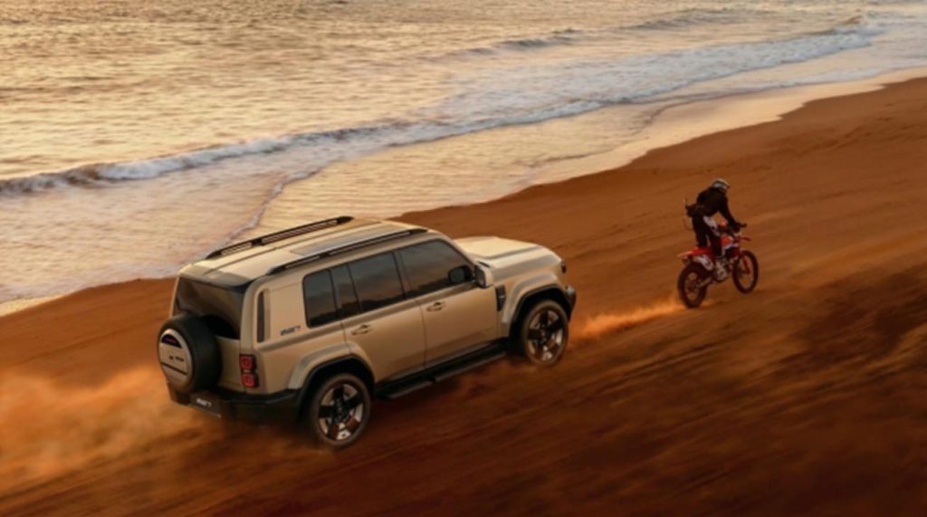 Un SUV iCAUR beige et une moto tout-terrain roulent sur le sable d'une plage au coucher du soleil, à côté de l'océan.