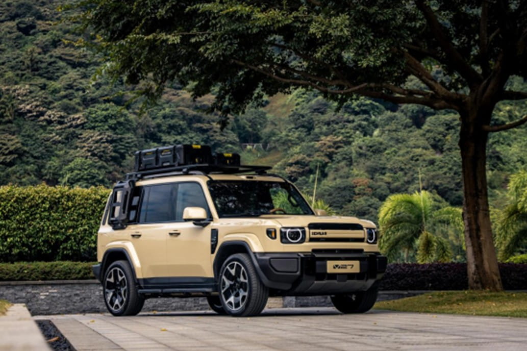 Photographie d'un SUV iCAUR beige, modèle V27, sur un parking pavé, avec une montagne verdoyante en arrière-plan.